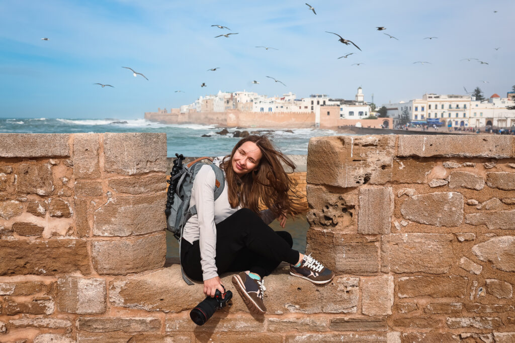 A girl with a camera sitting on a wall with a view of a old city and sea behind her 