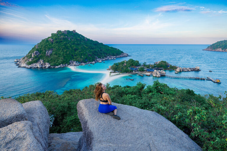 A girl half turned sitting on top of a boulder in focus with a view from Nang Yuan Viewpoint over an island